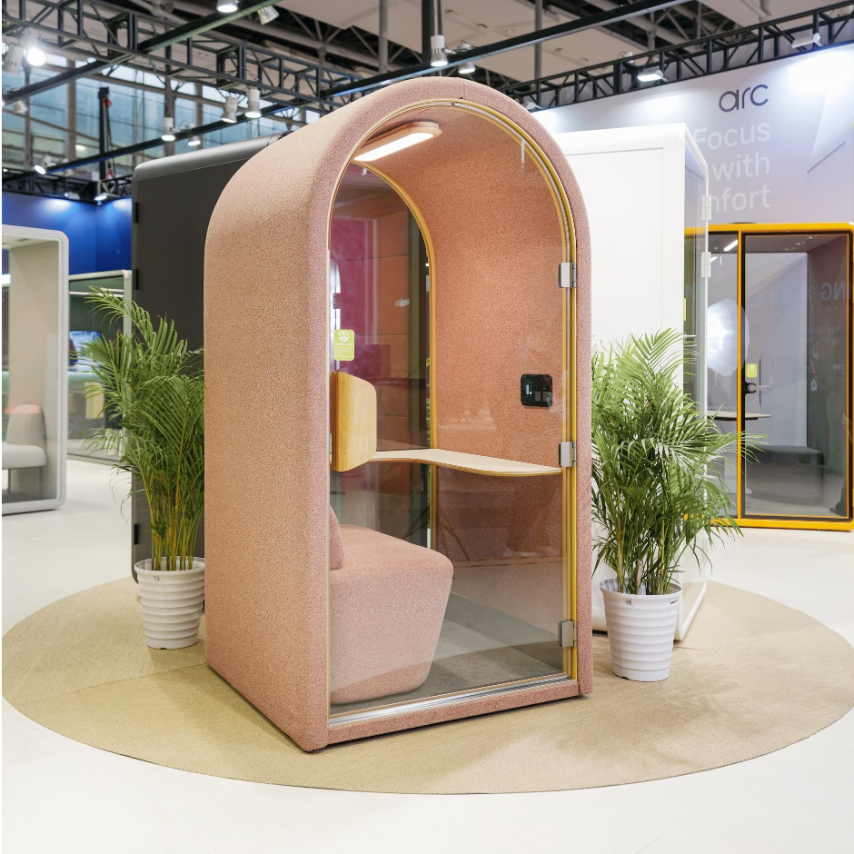 Blush pink acoustic pod with curved design, glass door, built-in desk and soft seating β ideal for quiet workspaces or private calls.