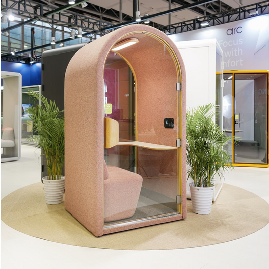 Blush pink acoustic pod with curved design, glass door, built-in desk and soft seating – ideal for quiet workspaces or private calls.